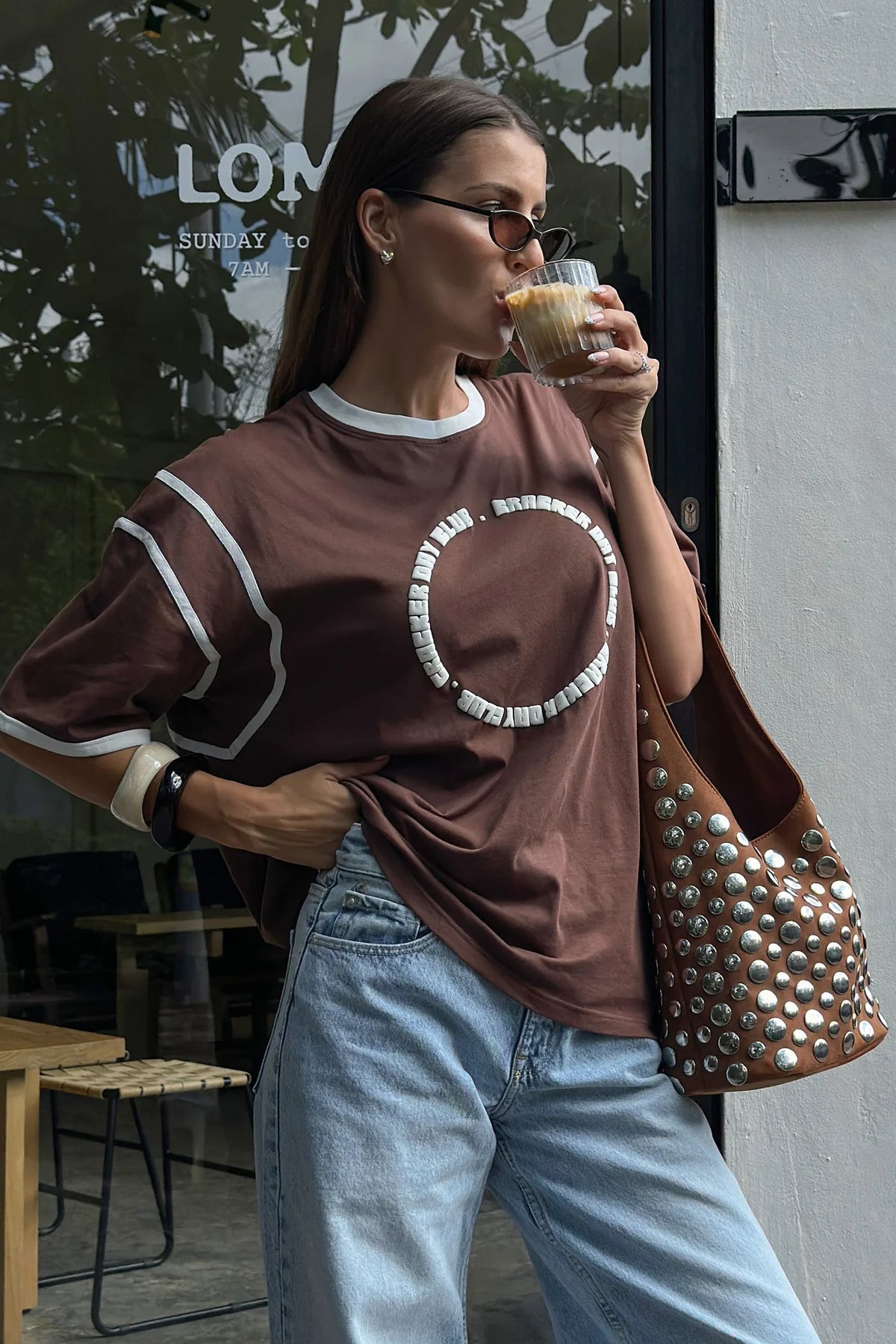 Woman drinking from a coffee cup outdoors, wearing a brown oversized t-shirt and blue jeans.
