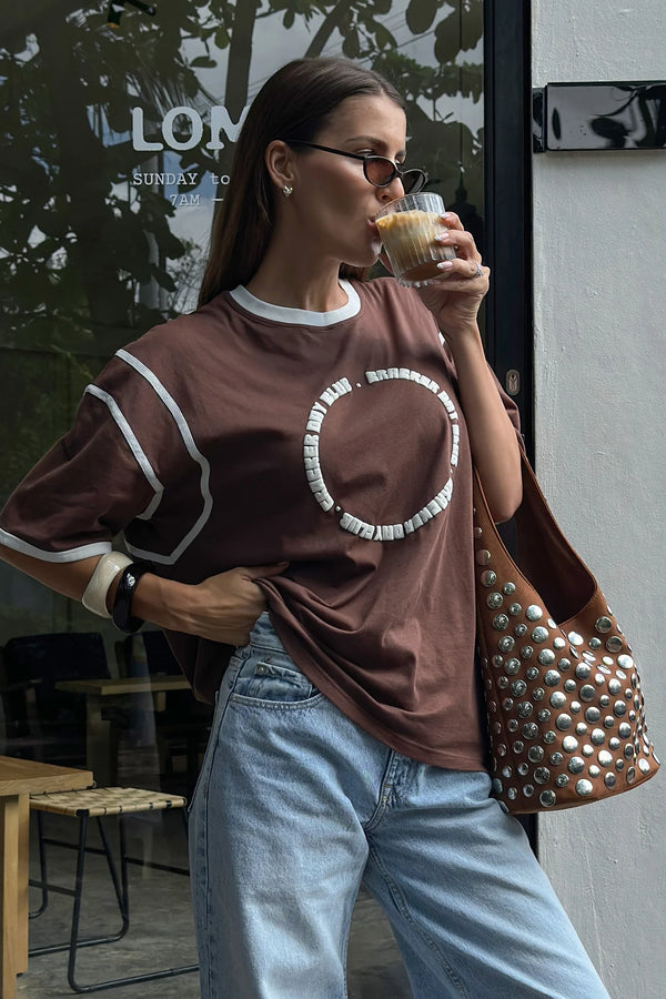 Woman drinking from a coffee cup outdoors, wearing a brown oversized t-shirt and blue jeans.