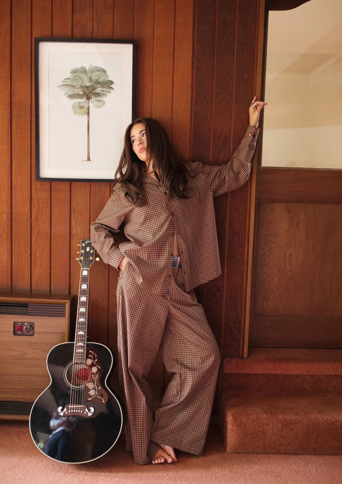 Woman in a FAR MER by Whitney ALL MY EXES plaid shirt and pants  outfit standing next to an acoustic guitar in a room with wooden paneling.