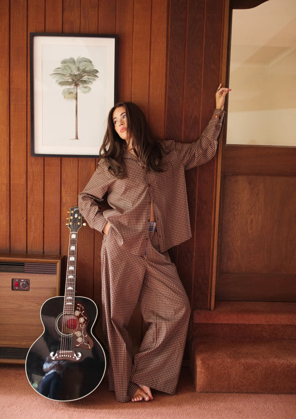 Woman in a FAR MER by Whitney ALL MY EXES plaid shirt and pants  outfit standing next to an acoustic guitar in a room with wooden paneling.