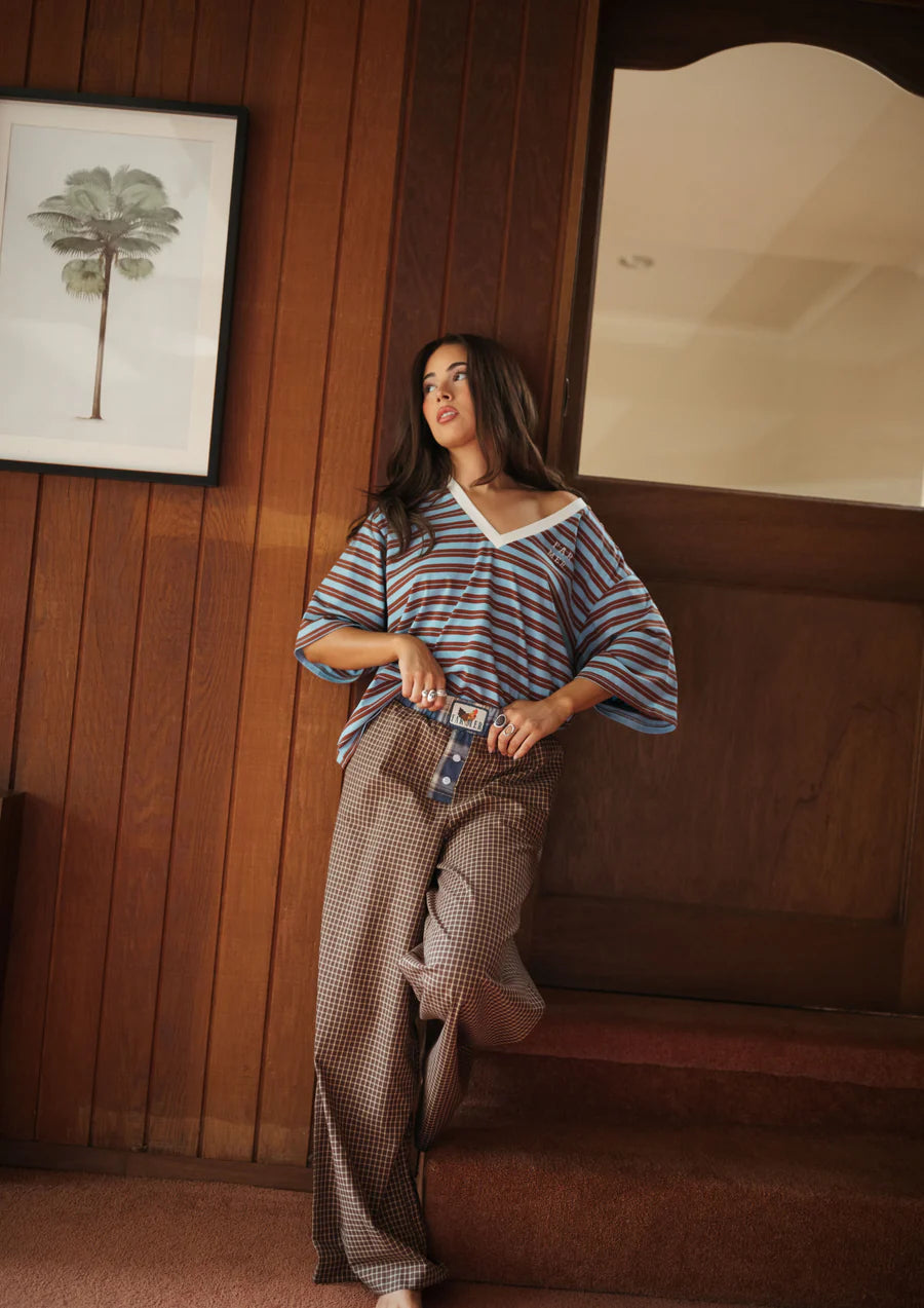 Woman standing wearing blue stripe tee and plaid pants from FAR MER by Whitney in a beige room leaning against a timber panel wall