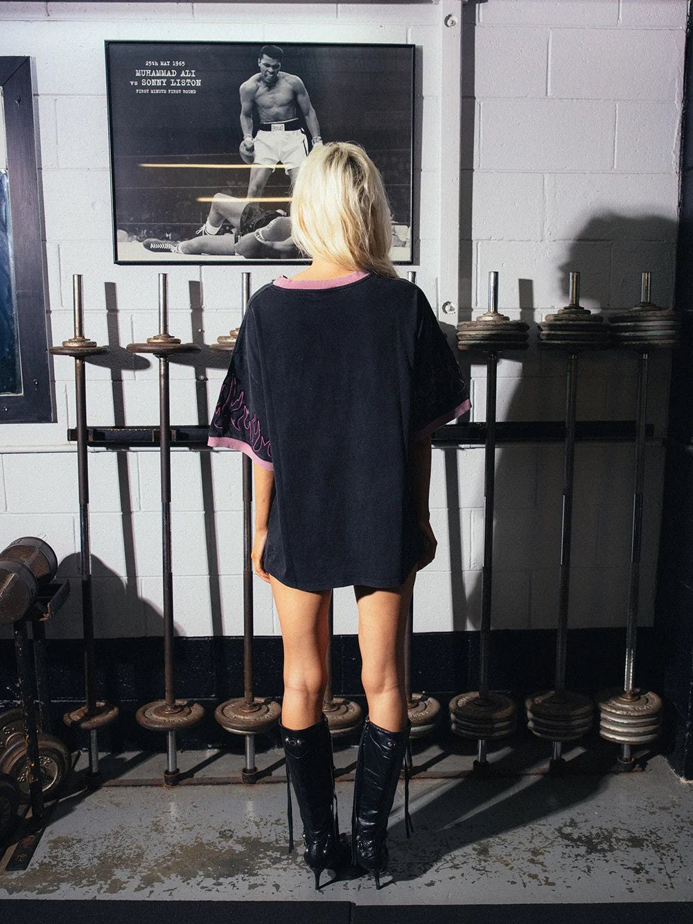 A woman stands with her back to the camera in a gym setting, wearing an oversized black tunic top with pink trim and black knee-high boots. She is positioned in front of a rack of swords and weight plates, with a framed boxing poster on the white brick wall behind her.