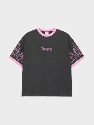 A black short-sleeve t-shirt with pink trim on the crew neck and sleeve cuffs, featuring pink flame-like graphic prints on the sleeves and a gothic-style 'justice' logo on the chest, displayed on a light gray background.