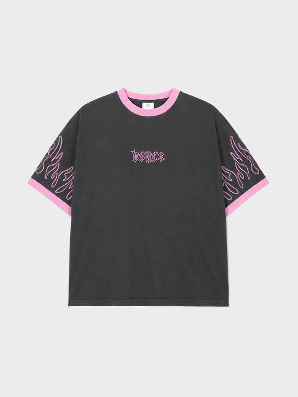 A black short-sleeve t-shirt with pink trim on the crew neck and sleeve cuffs, featuring pink flame-like graphic prints on the sleeves and a gothic-style 'justice' logo on the chest, displayed on a light gray background.