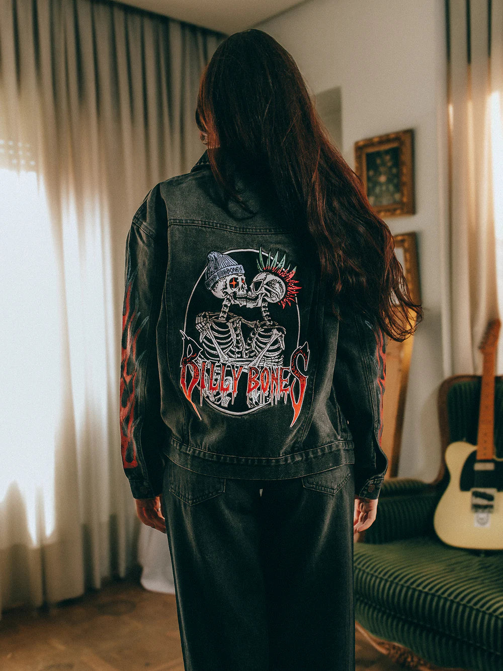 Person wearing a black denim jacket with a graphic design, standing in a room with curtains and a guitar.