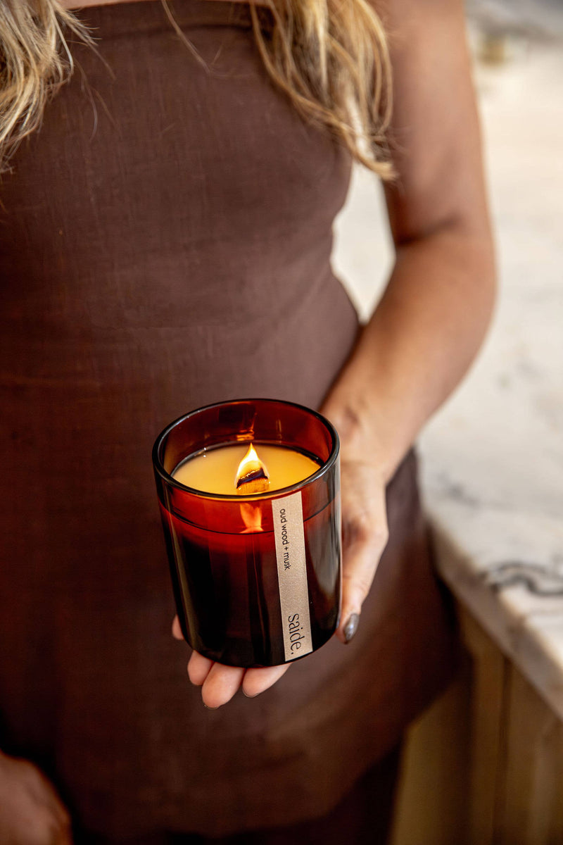 Person holding a lit candle in a dark container with 'SOM' branding.