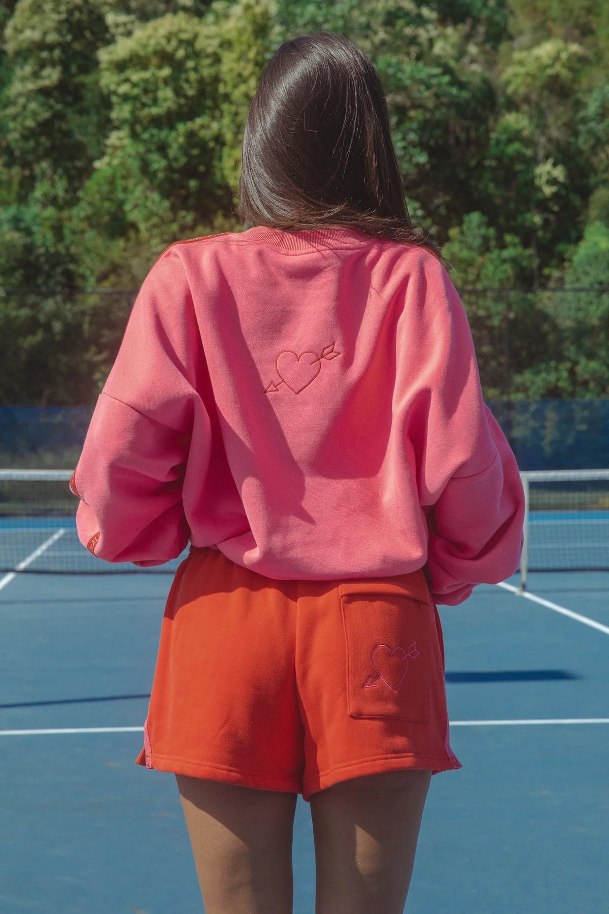 Person wearing a pink sweatshirt and orange shorts on a tennis court with trees in the background