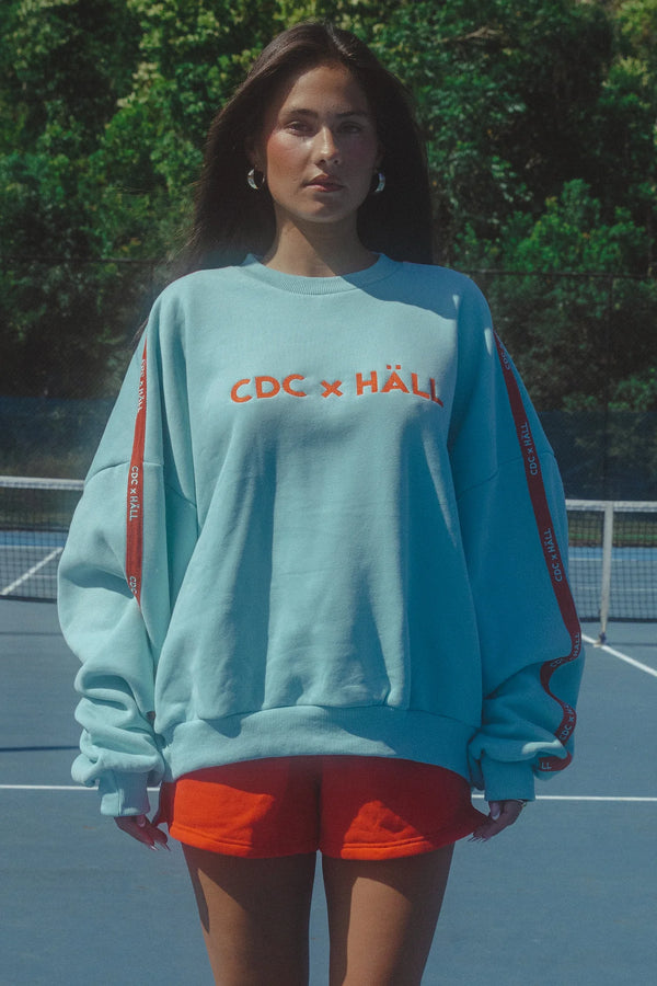 Person wearing a light blue sweatshirt with 'CDC x HAL' branding on a tennis court.