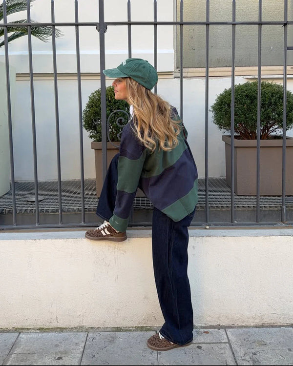 Person wearing a green cap, patterned sweater, and dark pants standing against a white wall with a metal fence and plants in the background.
