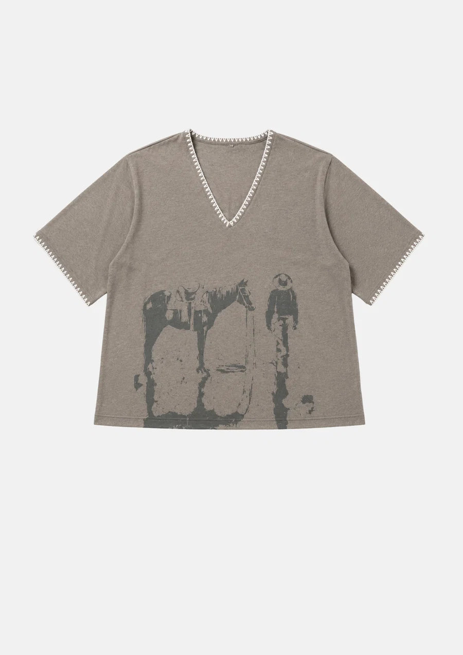 A taupe gray short-sleeve V-neck t-shirt with a distressed black graphic print of a cowboy riding a horse, featuring decorative white trim along the neckline and sleeve cuffs, shown flat on a light gray background.