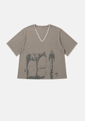 A taupe gray short-sleeve V-neck t-shirt with a distressed black graphic print of a cowboy riding a horse, featuring decorative white trim along the neckline and sleeve cuffs, shown flat on a light gray background.