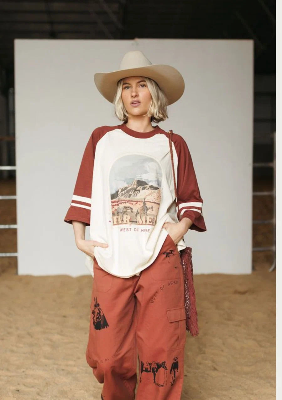 A woman modeling a western-themed raglan t-shirt with a graphic print, paired with rust-colored cargo pants and a beige cowboy hat, standing in front of a white studio backdrop.