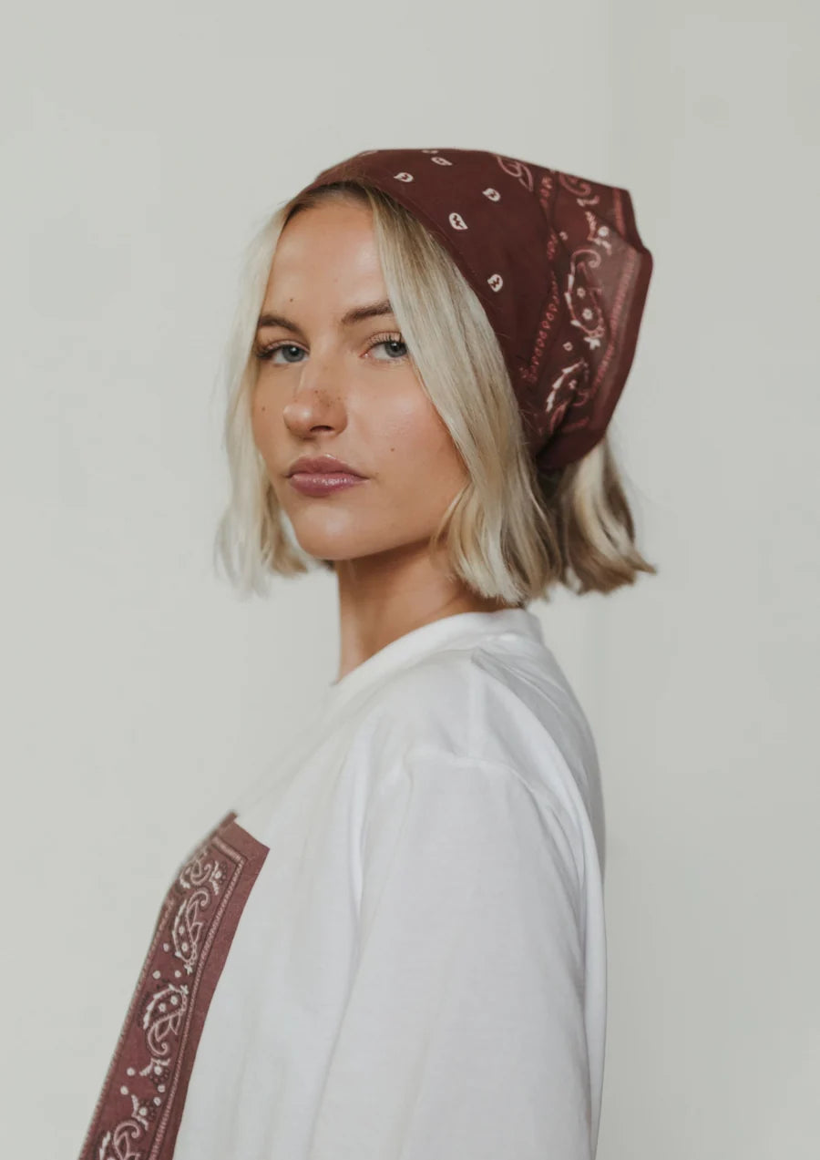 A woman with short blonde hair wearing a brown bandana with a white paisley pattern, styled as a headscarf, paired with a white t-shirt against a plain light background.