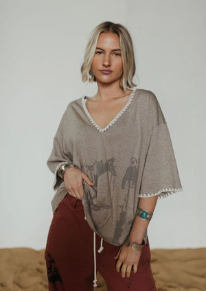 A woman modeling a loose taupe-gray V-neck top with white pom-pom trim and a horse graphic, paired with rust-colored pants, against a neutral studio backdrop.