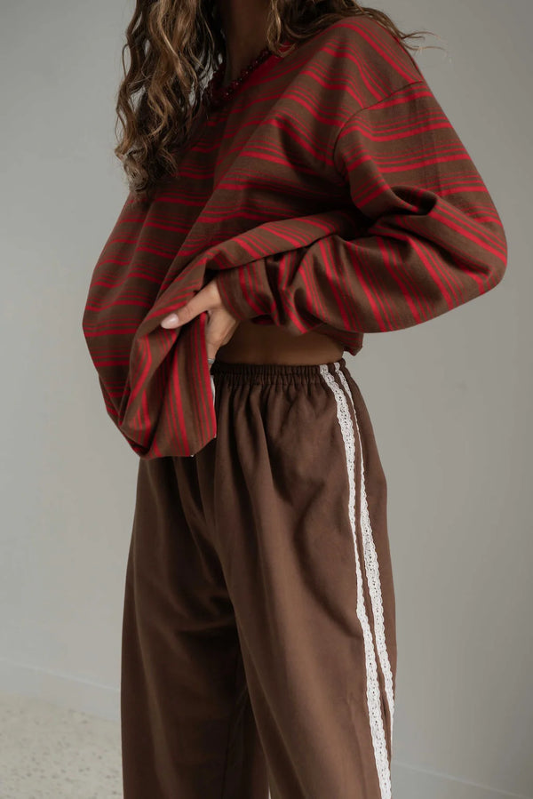 woman wearing brown pants with a white lace stripe and a brown and red striped long sleeve that she is holding up at the waist