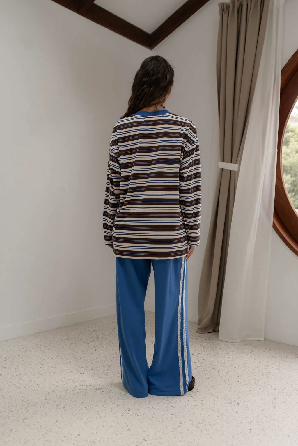 back view of woman near window wearing striped blue brown and oat long sleeve tee and blue pants