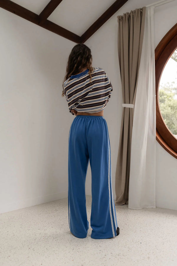 rear view of woman standing near window wearing brown white and blue stripe long sleeve tee and blue pants with white lace stripe