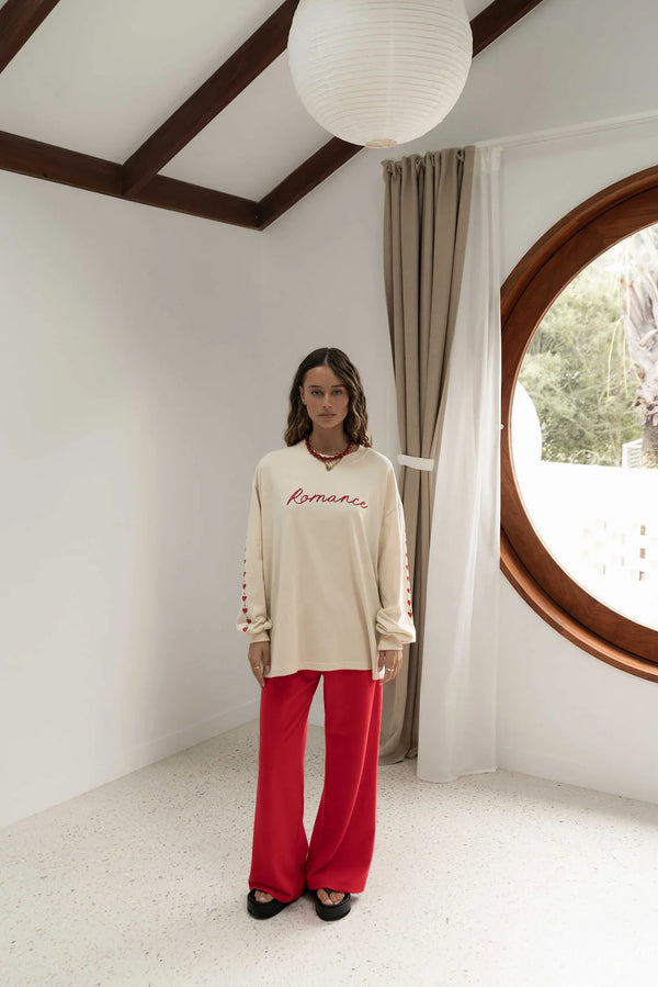 woman near a window wearing oat coloured long sleeve tee with romance written in red across the front and red pants