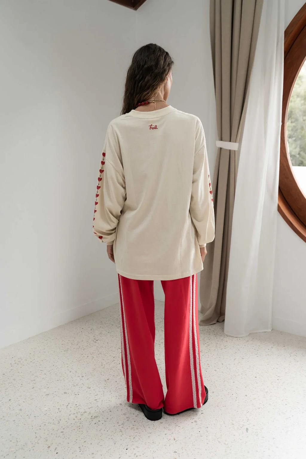 rear view woman near a window wearing oat coloured long sleeve tee with small Hall written in red at the neckline, red hearts down the sleeve and red pants