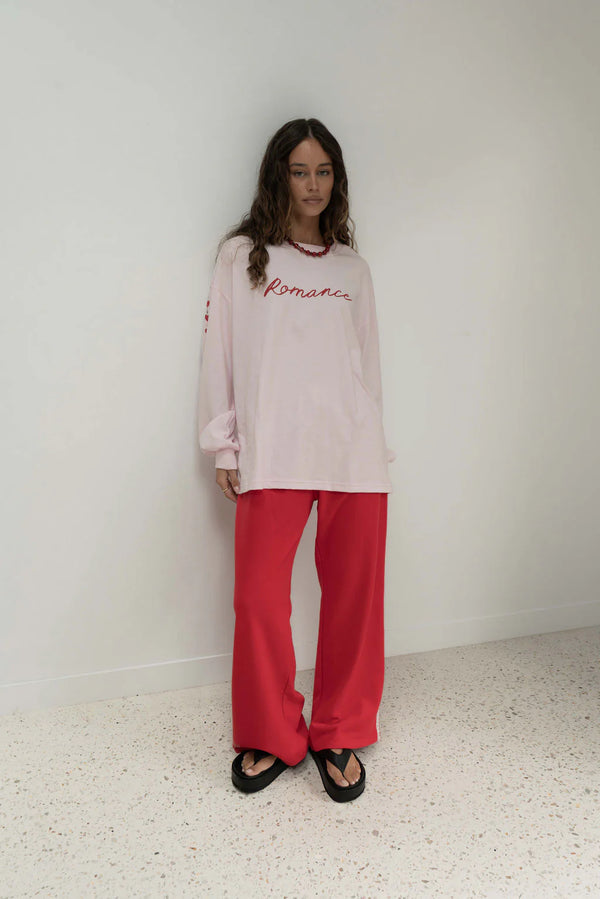 woman standing against white wall, wearing long sleeve pink tee with romance written across, red pants and black thongs