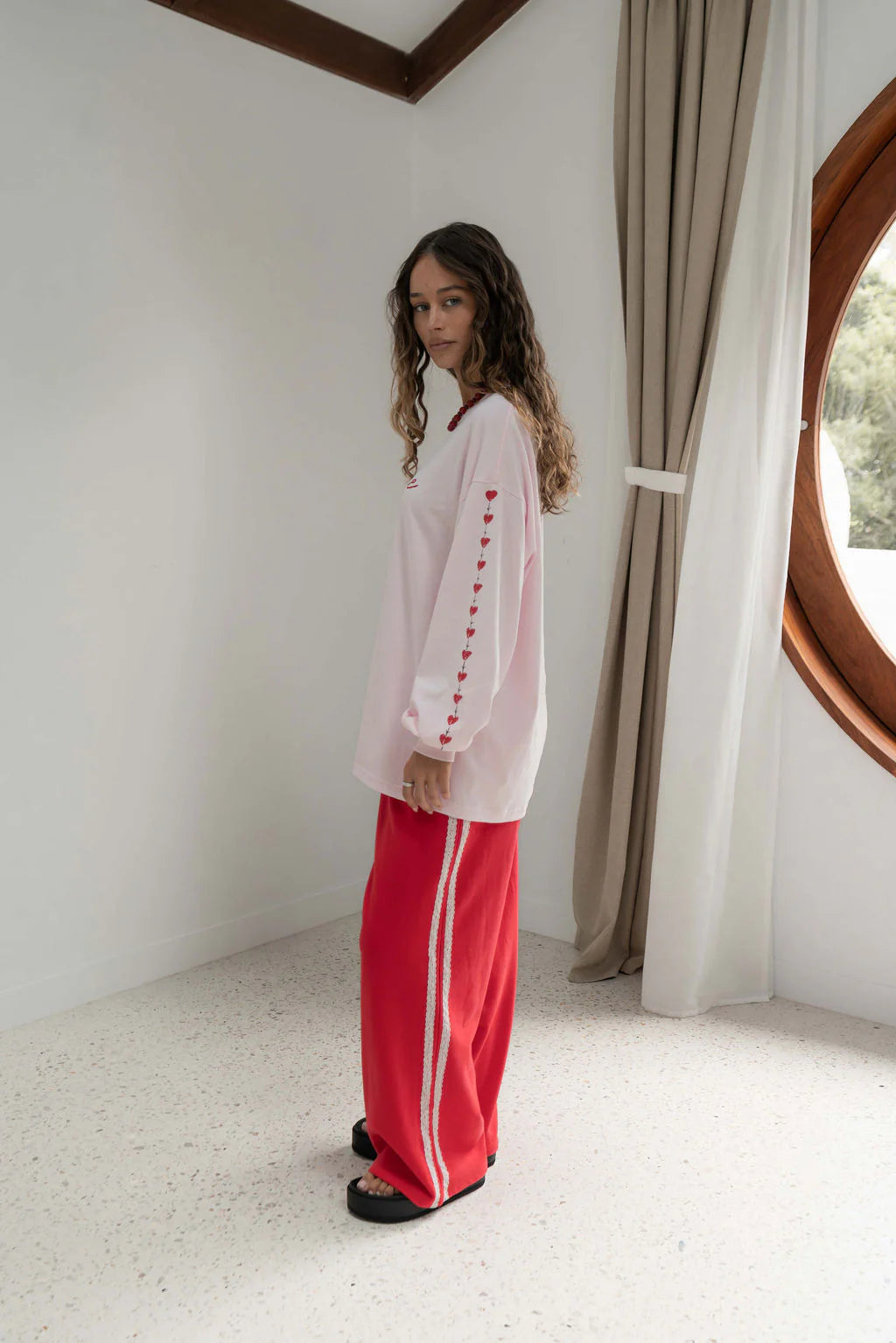 side view of woman standing in white bedroom, wearing pink long sleeve tee with love hearts down the arm, and red pants with a light pink stripe