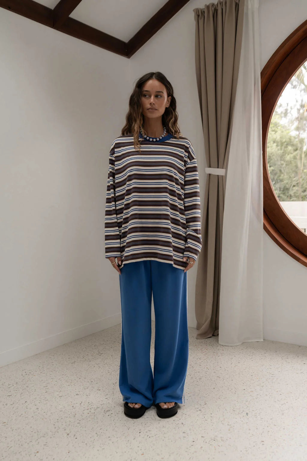 woman near window wearing striped blue brown and oat long sleeve tee and blue pants