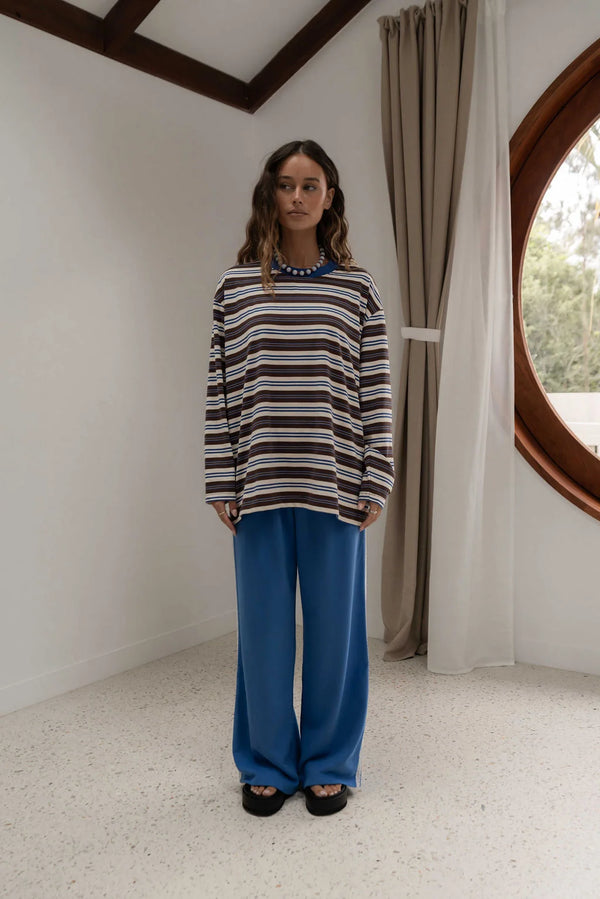 woman near window wearing striped blue brown and oat long sleeve tee and blue pants