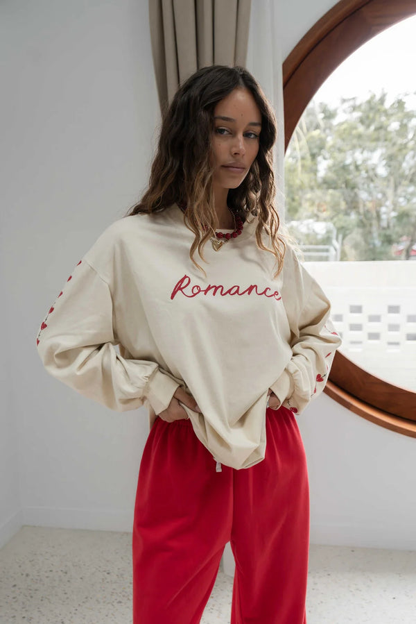woman near a window wearing oat coloured long sleeve tee with romance written in red across the front and red pants