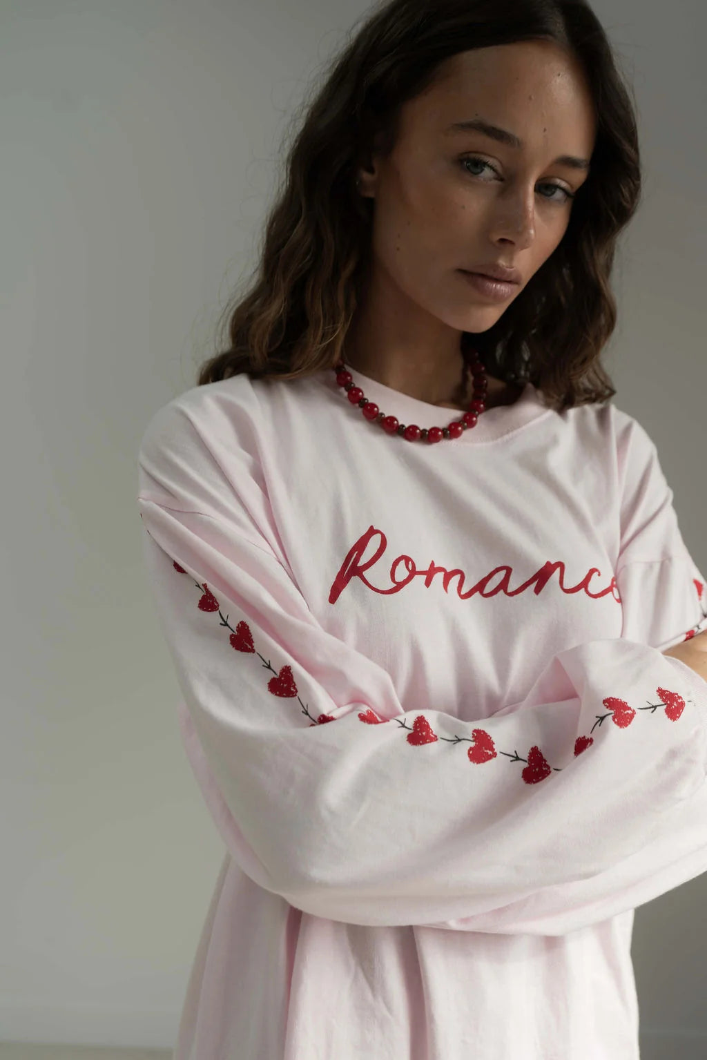 woman standing with arms crossed against white background, wearing pink long sleeve tee with romance in red written across chest and red hearts down the sleeve